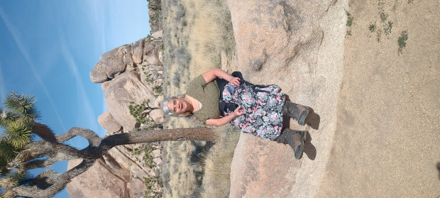 Deena Larsen in greenshirt and flowered skirt on a rock in front of a Joshua Tree and rock formation