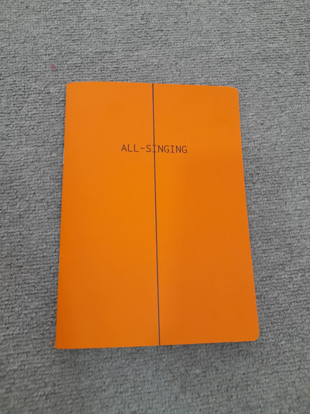 Photograph of "All-Singing"'s front cover. The book is bright orange, lying on a grey carpet.