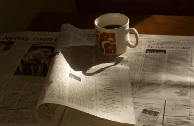 The first image seen in En anarchist er død is a photo of a newspaper, open to an obituary, with a coffee cup. The camera moves in closer.