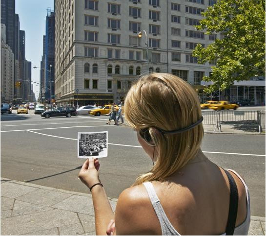 A woman holds up a historical photograph corresponding to the street she's walking on.