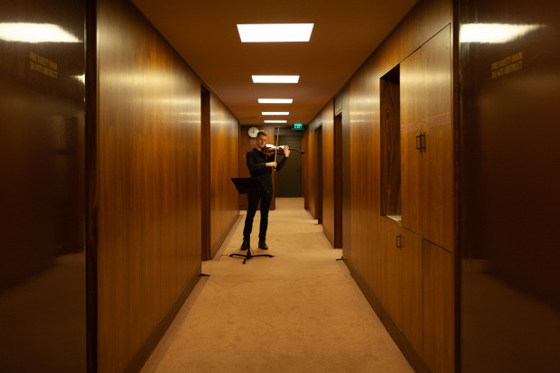 A man playing a violin in a long corridor, with many doorways inset into wooden walls.
