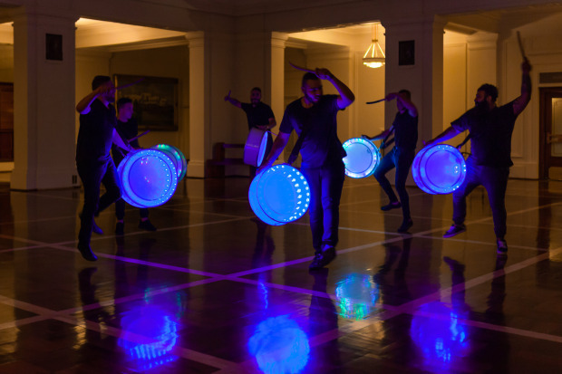 Six drummers play drums, illuminated with blue lights, in a dimly lit room with a polished floor.