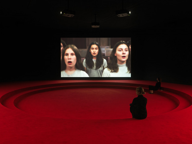 Two women sit in a large room with two raised ledges and red floor, watching "Assembly".