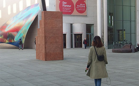 A woman with over-ear headphones walks away from the camera, outside of a modern looking museum.